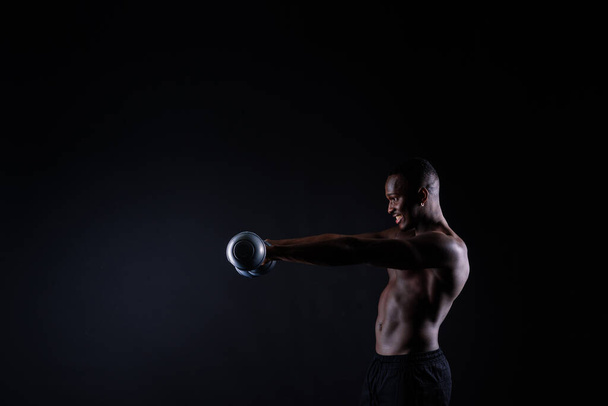 Απομονωμένος Αφρικανός μυώδης άντρας με αλτήρες σε σκοτεινό φόντο στούντιο. Δυνατός μαύρος χωρίς μπλούζα. - Φωτογραφία, εικόνα