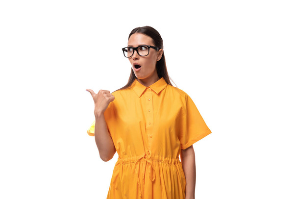europeo joven morena dama vestida con un vestido de verano naranja y gafas señala su dedo a un lado en sorpresa. - Foto, imagen