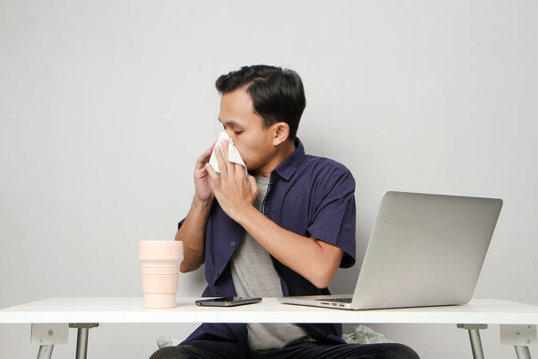У азиатского мужчины насморк, он вытирает нос салфеткой на рабочем месте, сидя перед ноутбуком. изолированный фон - Фото, изображение