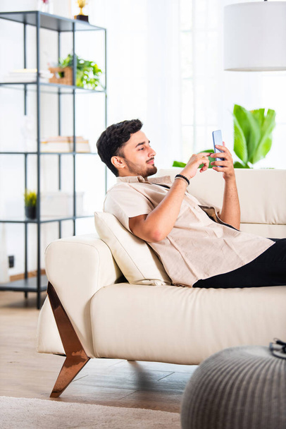 Mladý indický pohledný muž pomocí smartphone v obývacím pokoji, zatímco sedí na pohovce - Fotografie, Obrázek