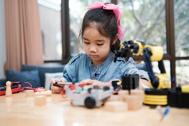 A young Asian girl in a STEM class attentively uses a smartphone app to remotely control a toy car, showcasing tech education. - Photo, Image