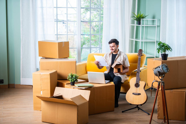 Індійський чоловік сидить на дивані з картонними коробками, шукає оренду нерухомості на ноутбуці або смартфоні - Фото, зображення