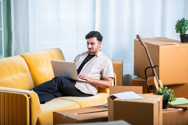 Uomo indiano seduto sul divano con scatole di cartone, cerca proprietà in affitto su laptop o smartphone - Foto, immagini