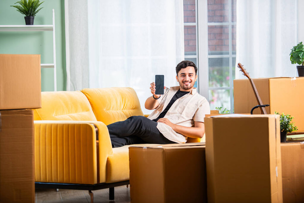 Индиец сидит на диване с картонными коробками, ищет арендуемую собственность на ноутбуке или смартфоне - Фото, изображение