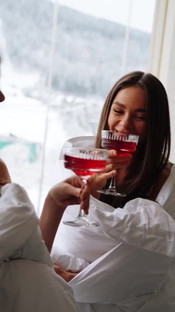 Dwie młode kobiety w kocach piją czerwone koktajle i bawią się na balkonie z widokiem na góry. Koncepcja wakacji hotelowych. Pionowe wideo - Materiał filmowy, wideo
