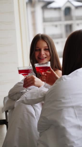 Kaksi nuorta naista huopissa juo punaisia cocktaileja ja pitää hauskaa parvekkeella näköalalla vuorille. Hotellin lomakonsepti. Pystysuora video - Materiaali, video