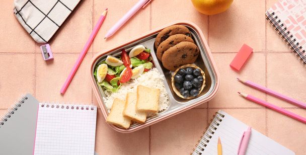 Scatola da pranzo con cibo gustoso e cancelleria su sfondo di piastrelle rosa - Foto, immagini