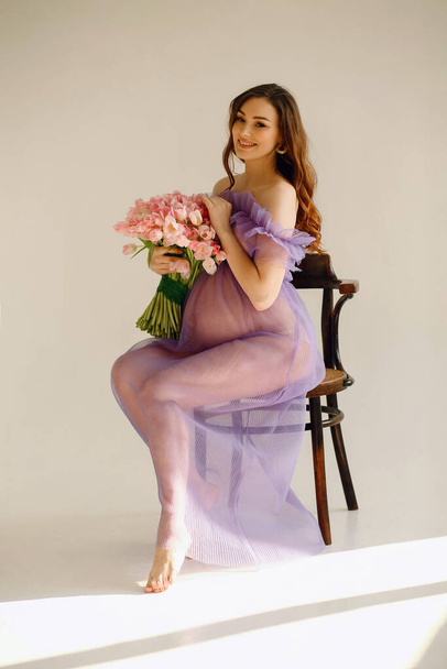 Attractive pregnant woman in violet dress sitting on the chair and holding a pink bouquet of tulips while posing in the studio on cyclorama. Stylish motherhood concept. Beautiful pregnancy - Photo, Image