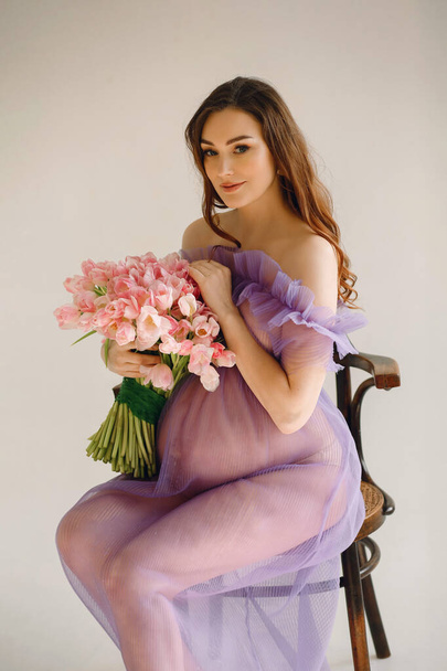 Портрет привабливої вагітної жінки в фіолетовій сукні, що сидить на стільці і тримає рожевий букет тюльпанів, позуючи в студії на білому тлі. Стильна концепція материнства. Красива вагітність - Фото, зображення