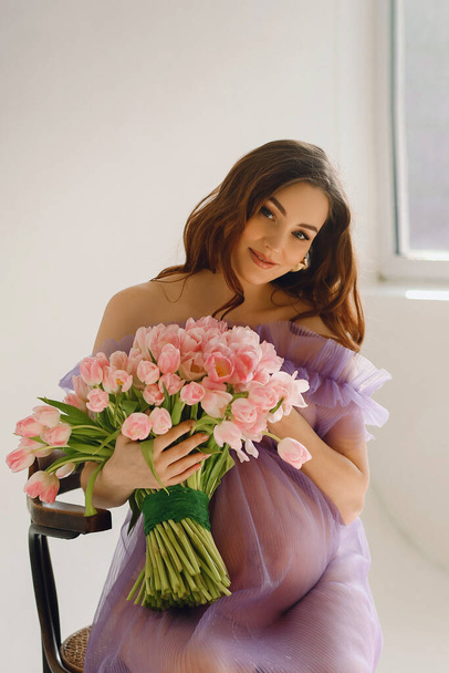 Aantrekkelijke zwangere vrouw in violette jurk zittend op de stoel en een roze boeket tulpen vasthoudend terwijl ze poseert in de studio. Stijlvol moederschap concept. Mooie zwangerschap - Foto, afbeelding