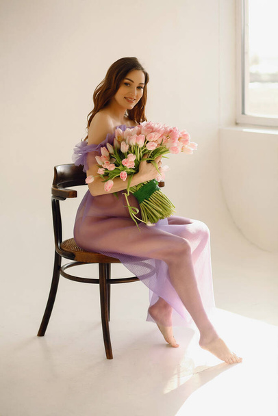 Attraktive schwangere Frau in violettem Kleid sitzt auf dem Stuhl und hält einen rosafarbenen Tulpenstrauß in der Hand, während sie im Studio posiert. Glückliche Mutterschaft Konzept. Schöne Schwangerschaft - Foto, Bild
