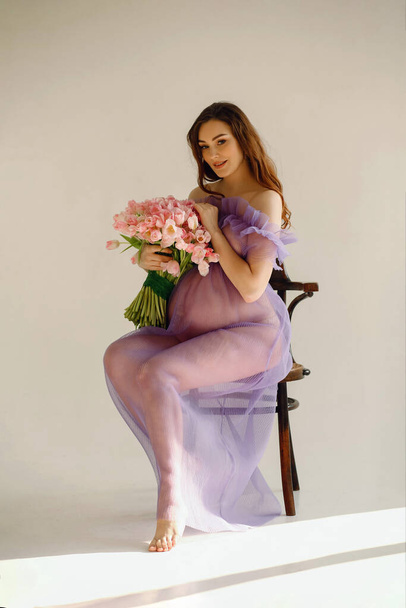 Приваблива вагітна жінка в фіолетовій сукні сидить на стільці і тримає рожевий букет тюльпанів, позуючи в студії на білому тлі. Стильна концепція материнства. Красива вагітність - Фото, зображення