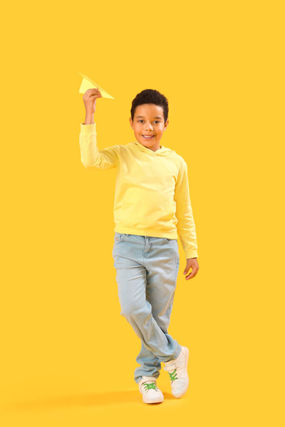 Ένα Αφροαμερικανάκι με χάρτινο αεροπλάνο σε κίτρινο φόντο. Γιορτή της ημέρας των παιδιών - Φωτογραφία, εικόνα