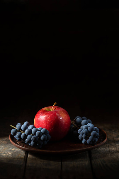 uvas azuis e maçã vermelha em uma chapa de barro em uma mesa de madeira - Foto, Imagem
