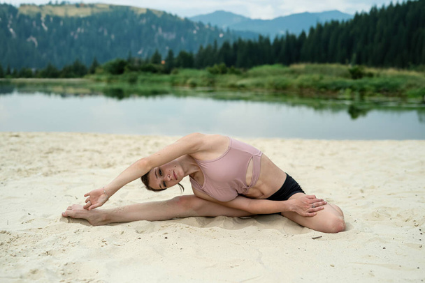 Молодая женщина практикует йогу на песчаном пляже возле озера с горным фоном. Стретч упражнения на природе солнечный день - Фото, изображение