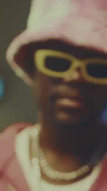 Vertical zoom-in closeup portrait of stylish African American hipster guy posing for camera indoors wearing pink plushy bucket hat and yellow glasses - Footage, Video