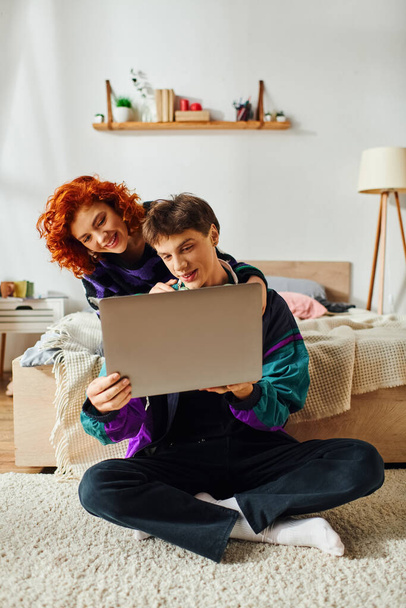 σαγηνευτικό χαρούμενο νεαρό ζευγάρι με ζωντανά κομψά ρούχα που περνούν το χρόνο τους στο φορητό υπολογιστή μαζί στο σπίτι - Φωτογραφία, εικόνα