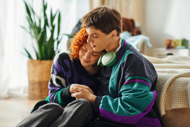 hombre guapo con auriculares abrazando amorosamente a su hermosa novia pelirroja mientras está en casa - Foto, Imagen
