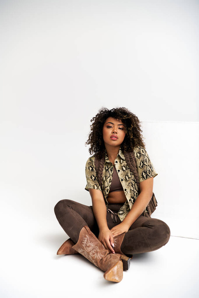 привлекательная африканская американка с отпечатками на одежде, сидящая на полу и смотрящая в камеру - Фото, изображение