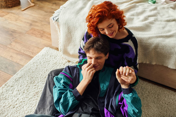 passionate red haired woman in cozy attire smiling and hugging her loving handsome boyfriend - Photo, Image
