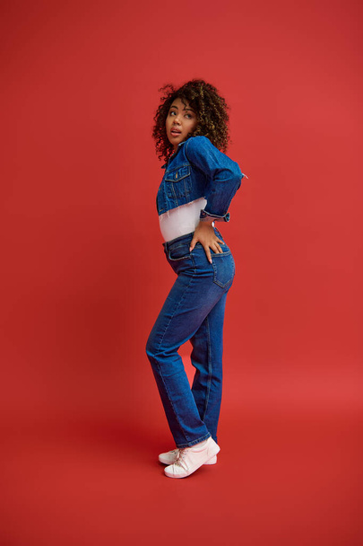 изящная молодая африканская американка в стильной джинсовой одежде, смотрящая на красный фон - Фото, изображение