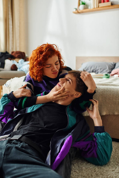 beautiful red haired woman in vibrant outfit hugging her loving joyous boyfriend while at home - Photo, Image
