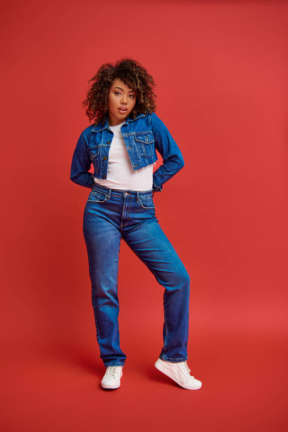 attractive chic african american woman in denim attire posing on red backdrop and looking at camera - Foto, immagini