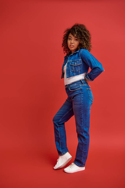fashionable african american woman in denim attire posing on red backdrop and looking at camera - Photo, Image