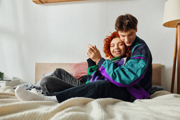 Χαρούμενος όμορφος άντρας με ακουστικά που αγκαλιάζει την κοπέλα του στοργικά ενώ είναι στο κρεβάτι - Φωτογραφία, εικόνα