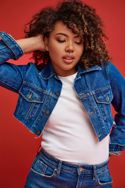 сложная молодая африканская американка в стильном джинсовом наряде смотрит в сторону на красном фоне - Фото, изображение
