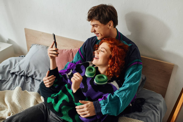 bel homme joyeux avec écouteurs modernes regardant le téléphone avec sa petite amie joyeuse sur le lit - Photo, image