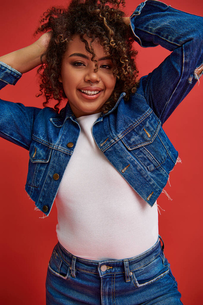 cheerful african american woman in denim attire posing on red backdrop and looking at camera - Photo, Image
