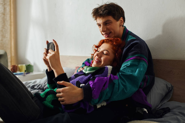 handsome joyous man with headphones using smartphone with his loving red haired girlfriend on bed - Photo, Image