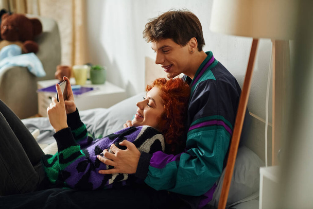 aantrekkelijke vrolijke man met koptelefoon kijken naar telefoon met zijn mooie vrolijke vriendin - Foto, afbeelding