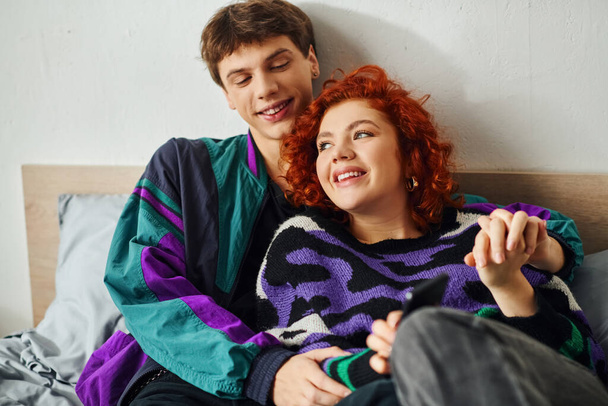joyous good looking couple in vibrant stylish attires hugging and laughing while in bed at home - Photo, Image
