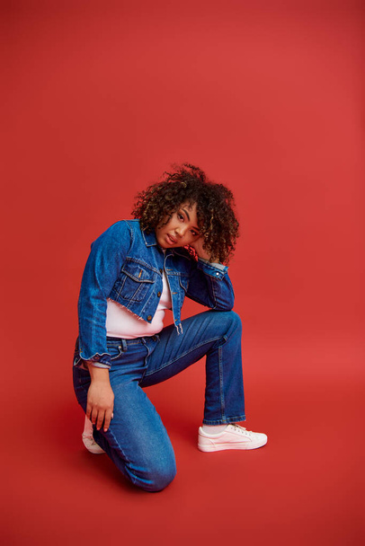 trendy african american fashionista in denim attire posing on red backdrop and looking at camera - Photo, Image