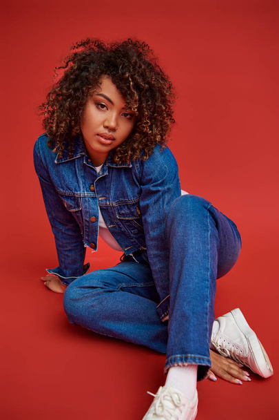 chic african american female model in denim attire looking at camera and posing on red background - Photo, Image