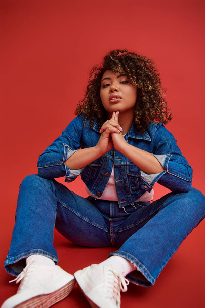 alluring african american female model in denim attire looking at camera and posing on red backdrop - Photo, Image