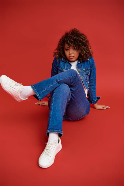 заманчивая африканская американская модель в джинсовой одежде, смотрящая в камеру и позируя на красном фоне - Фото, изображение