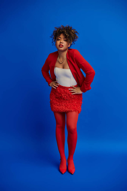 stílusos afro-amerikai nő piros szoknya és blézer pózol kék háttér és nézi a kamera - Fotó, kép