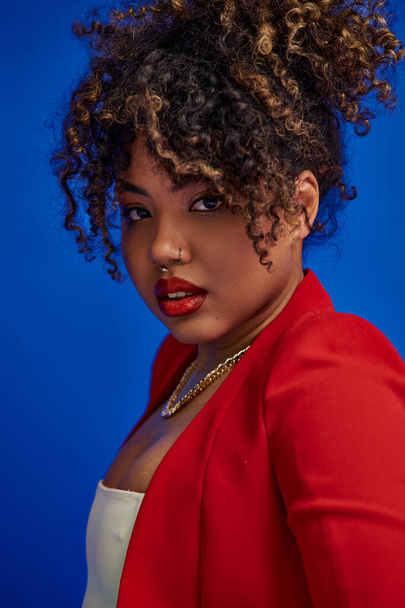 young african american woman in red skirt and blazer posing on blue backdrop and looking at camera - Foto, afbeelding