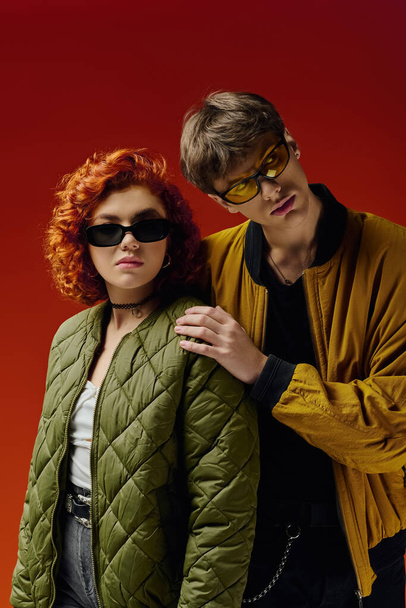 přitažlivý módní pár s módními slunečními brýlemi v útulných šatech pózuje na červeném pozadí spolu - Fotografie, Obrázek