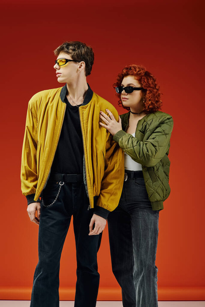 любящая пара в модных солнцезащитных очках в модных одеждах, позирующих вместе на красном фоне - Фото, изображение