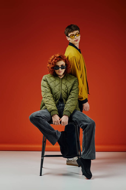 beautiful woman in sunglasses sitting on chair next to her fashionable boyfriend on red backdrop - Photo, Image