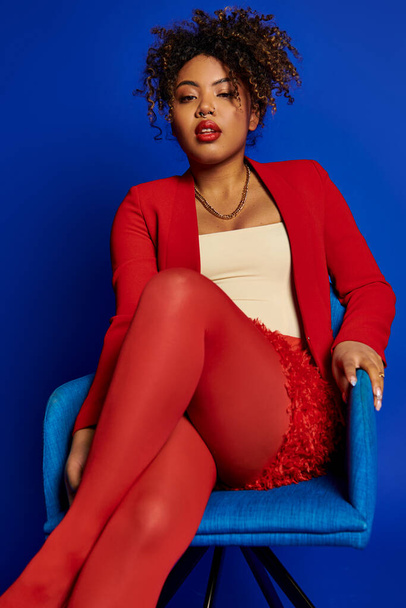 encantadora mujer afroamericana elegante en traje rojo inteligente sentado en la silla azul y mirando a la cámara - Foto, Imagen