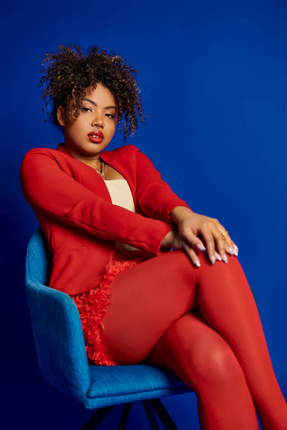 okouzlující elegantní africká americká žena v elegantním červeném obleku sedí na modré židli a dívá se do kamery - Fotografie, Obrázek