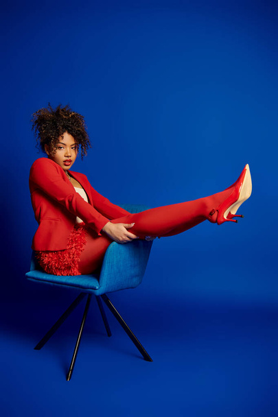 csábító elegáns afro-amerikai nő okos piros ruha ül a kék szék és a kamera - Fotó, kép