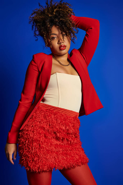 refined chic african american woman in red blazer with curly hair looking at camera, blue backdrop - Photo, Image