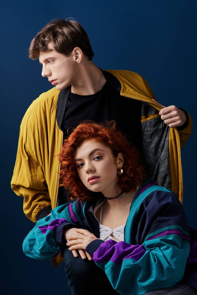 young woman in vivid attire looking at camera next to her handsome boyfriend on dark blue backdrop - Photo, Image