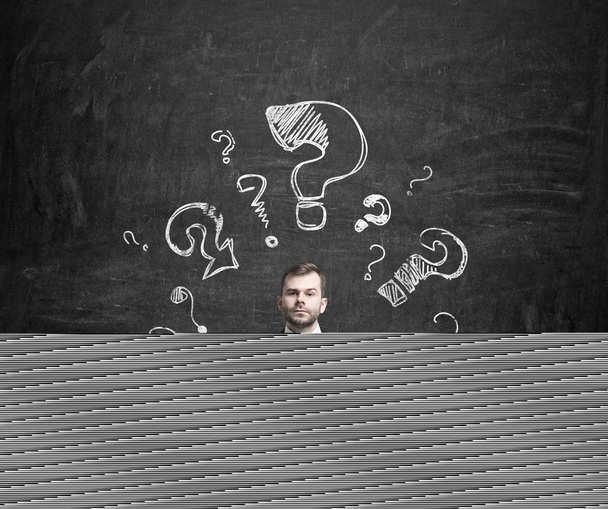 Young businessman is thinking about complicated questions. Drawn question marks on the dark wall. - Photo, Image